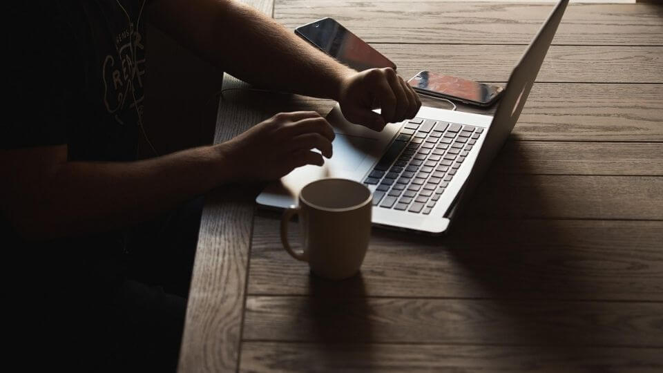 A person using a latop with a cup and two phones beside them.