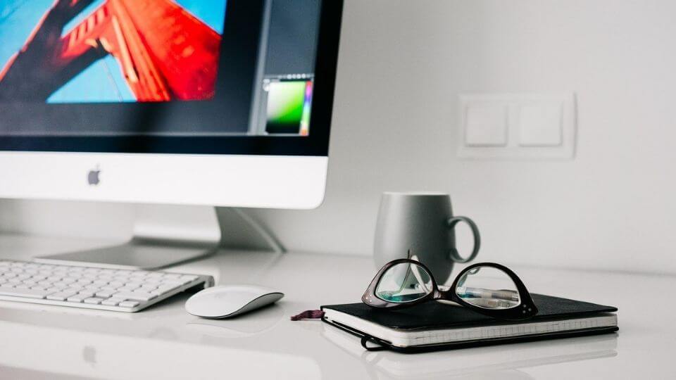 A workstation with a computer, keyboard, mouse, book, glasses, and mug.