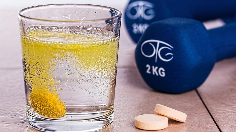 An effervescent vitamin in a glass with extra vitamins and dumbbells next to it.