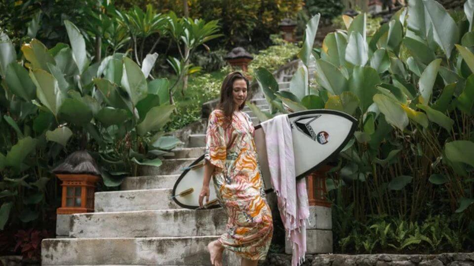 A person walks down steps holding a surf board and surrounded by gorgeous greenery at Pelan Pelan Surf Camp & Yoga Retreat in Bali.