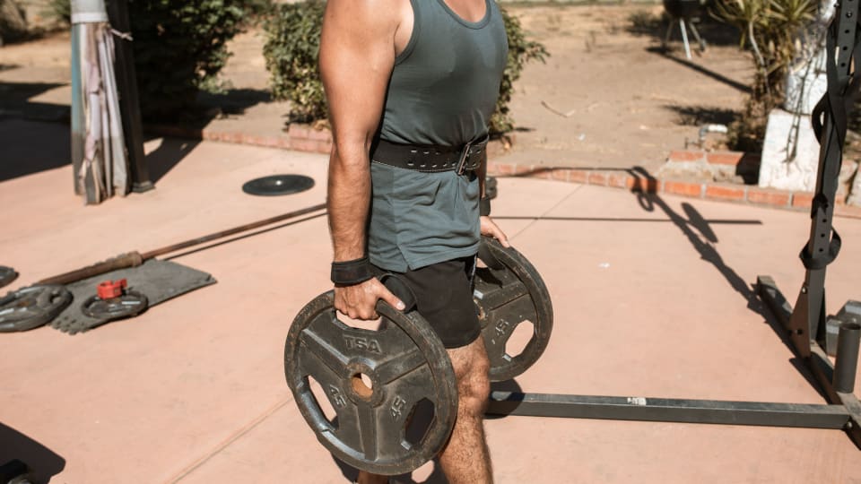 A person carries a weight plate in each hand in an outdoor gym setting.