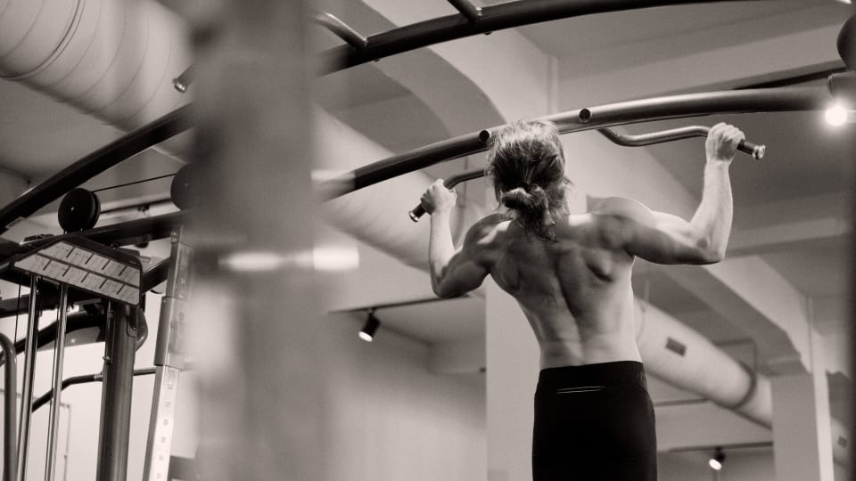A person doing a wide grip overhand pull up on a bar in a gym.