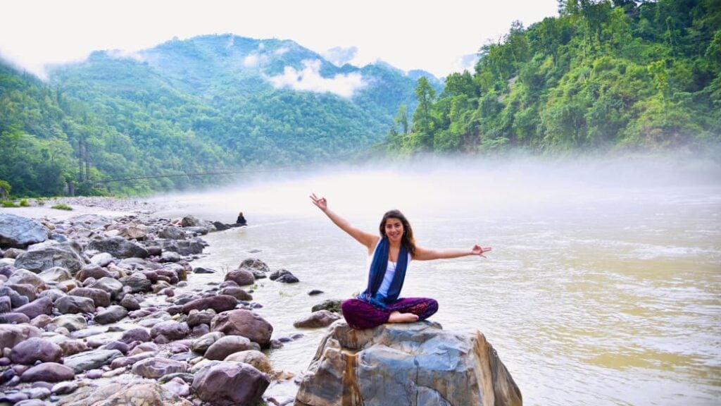 A happy person sits with legs crossed and arms stretched out on a rock in nature, surrounded by water and greenery.