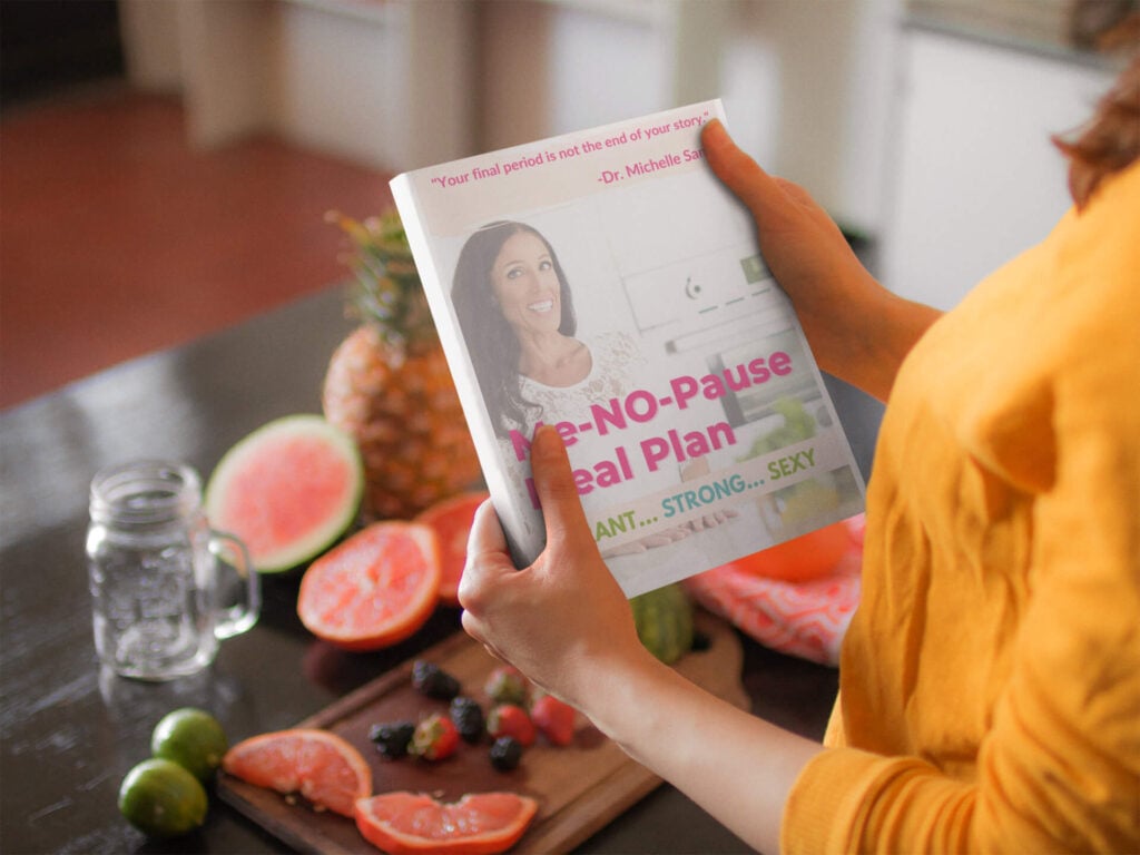A person holds Dr Michelle Sands Me-No_Pause meal plan guide, with chopped up fruit on the bench in the background.