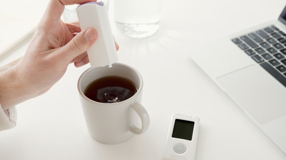 A person's hand adds a sweetening capsule from a container into a cup of coffee, with a laptop in the background.