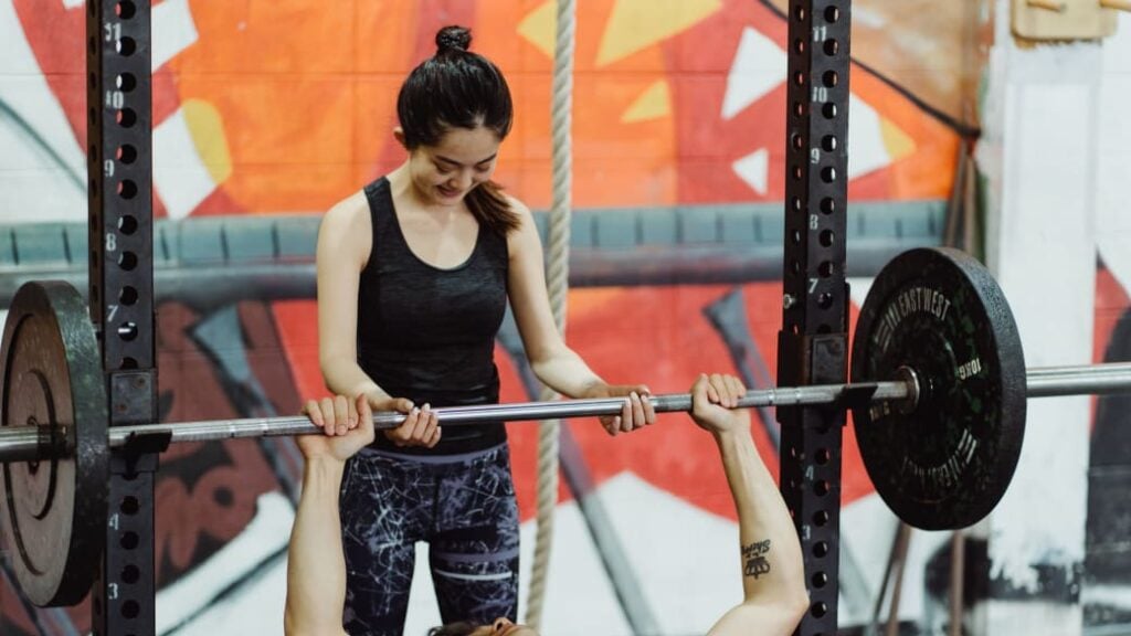 One person spots another for a bench press in a rack.