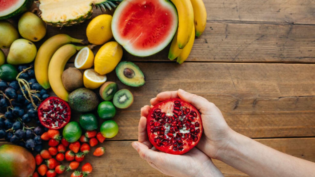 A cut pomegranate in a person's hands, with a colorful selection of fruit on a wooden table underneath.