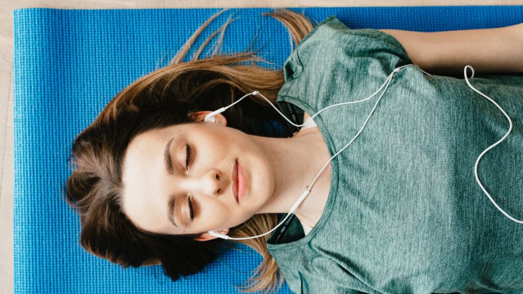 A person lies on a yoga mat on the ground, with eyes closed and earphones in, listening to a guided meditation.