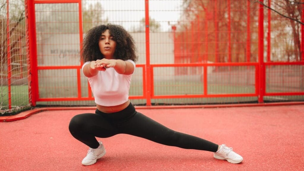 A person shows excellent hip mobility with a deep side lunge on a red outdoor court.