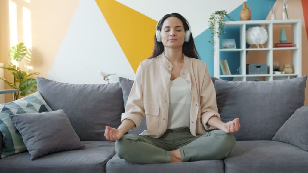 A person sits cross-legged on a sofa, with eyes closed, listening to a guided meditation with headphones on.
