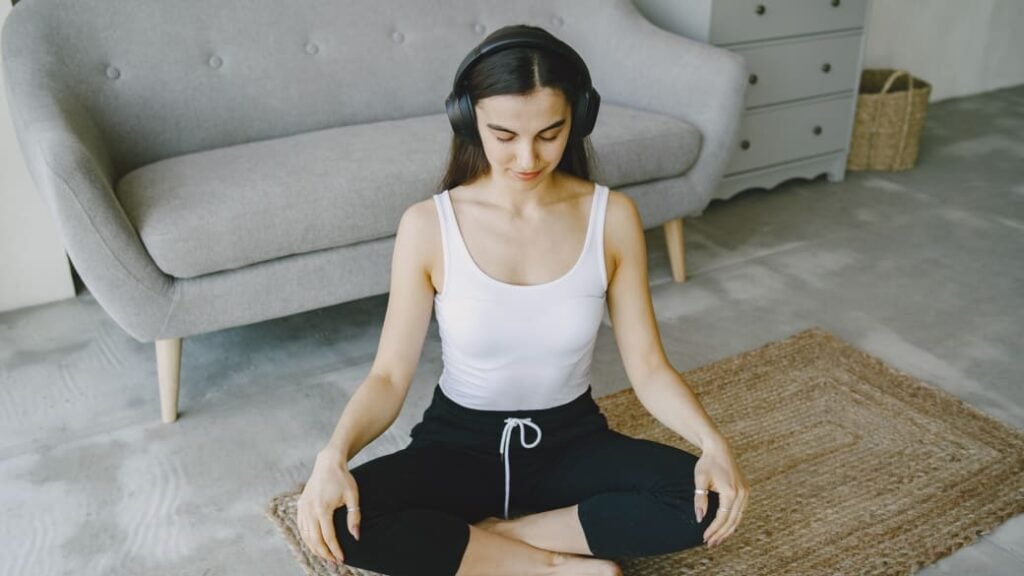 A person sits cross-legged on the floor with eyes closed and headphones on, appearing to listen to a guided meditation.